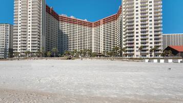 Um hotel grande com vários andares e varandas, uma praia arenosa em primeiro plano e palmeiras.