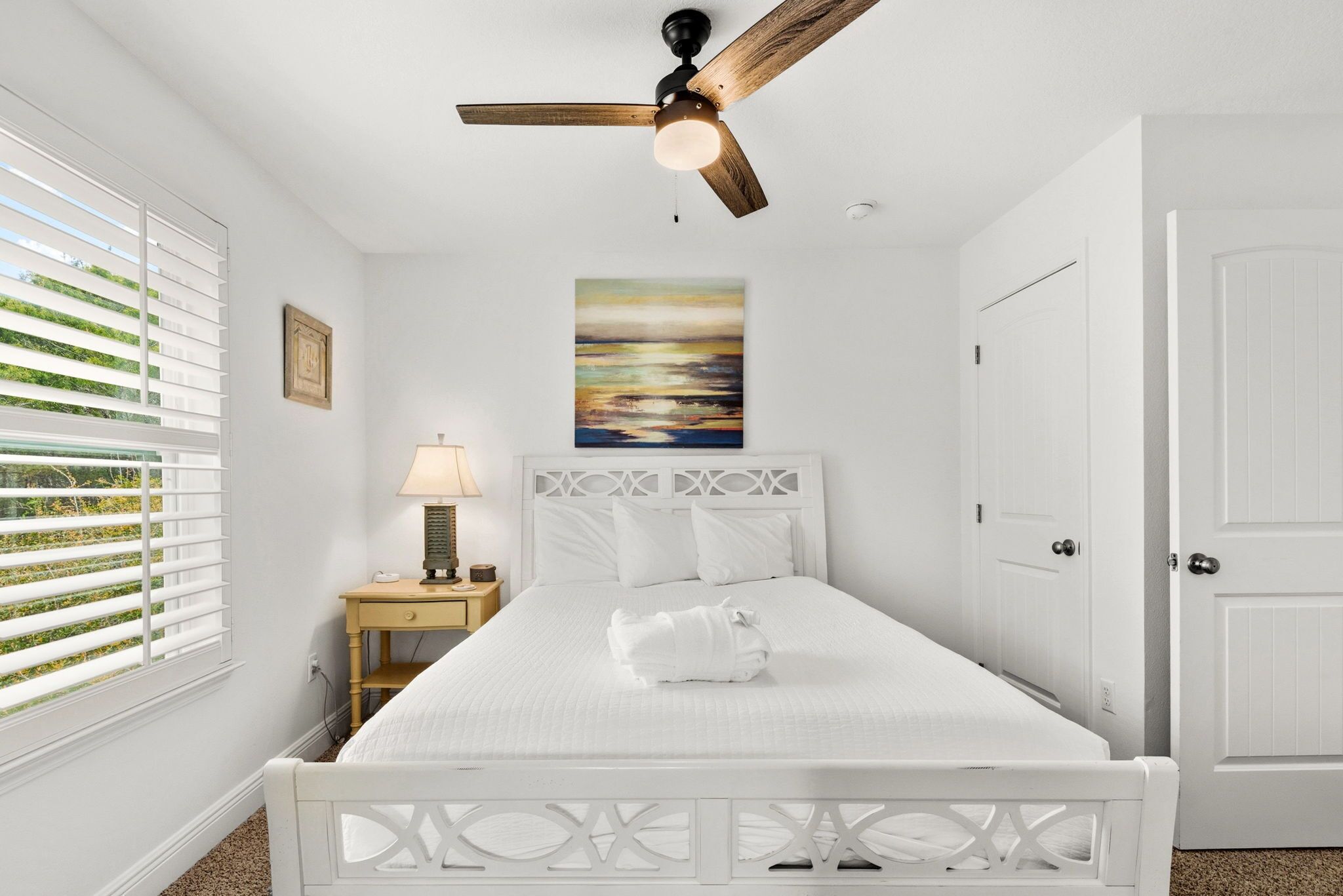 Une chambre à coucher blanche, avec un lit, une table de chevet, une lampe, un ventilateur de plafond, un tableau et une porte.