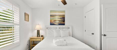 Une chambre à coucher blanche, avec un lit, une table de chevet, une lampe, un ventilateur de plafond, un tableau et une porte.
