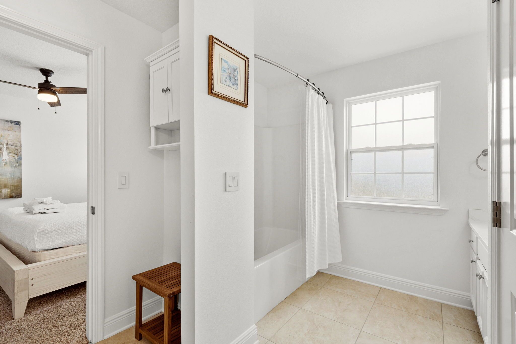 A bedroom with a bed, a ceiling fan, a small wooden table, and a bathroom with a shower curtain, a window, and a bathtub.