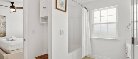 A bedroom with a bed, a ceiling fan, a small wooden table, and a bathroom with a shower curtain, a window, and a bathtub.