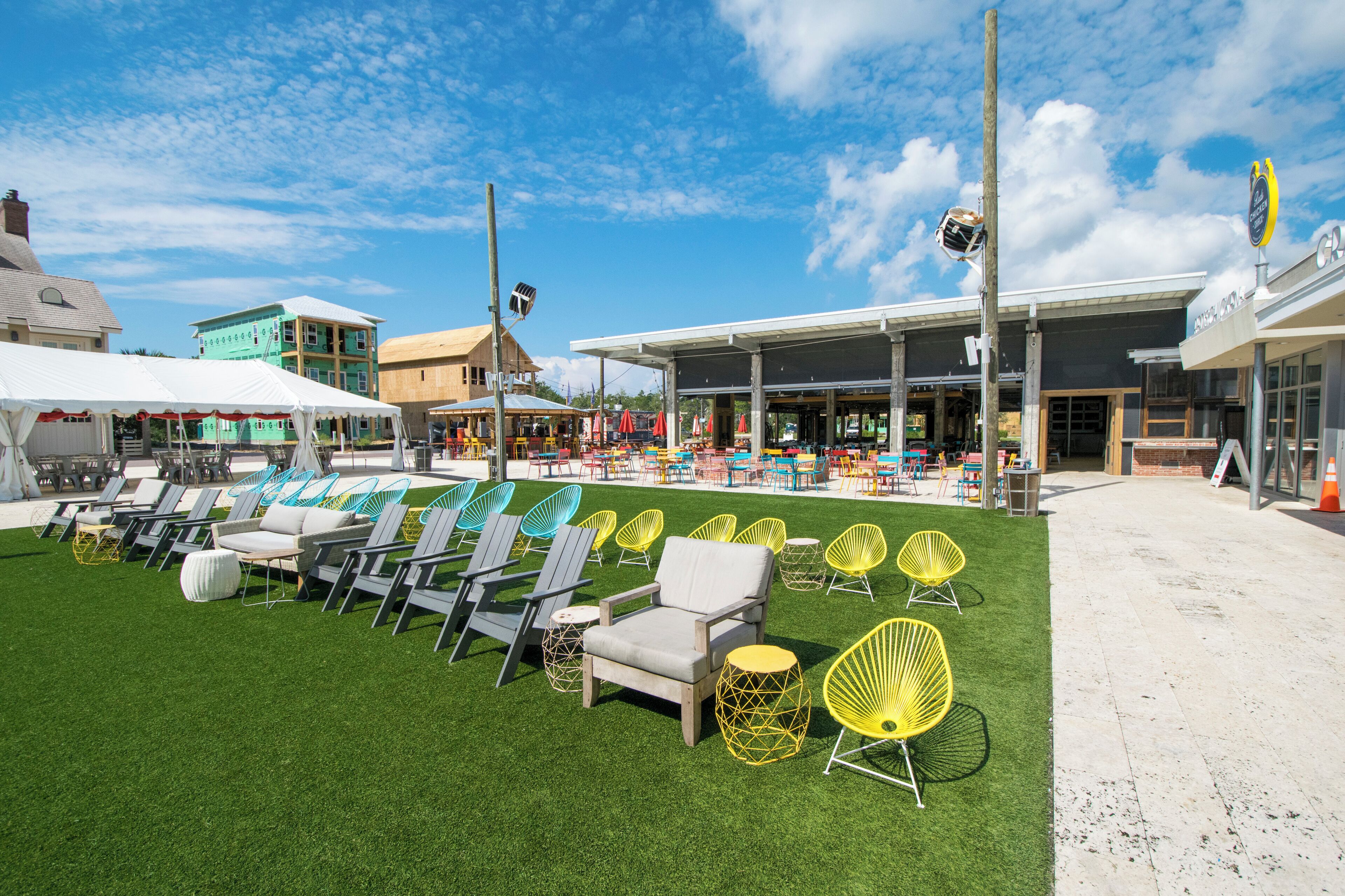 An outdoor seating area with rows of chairs and tables, a building with a storefront, and a grassy lawn.