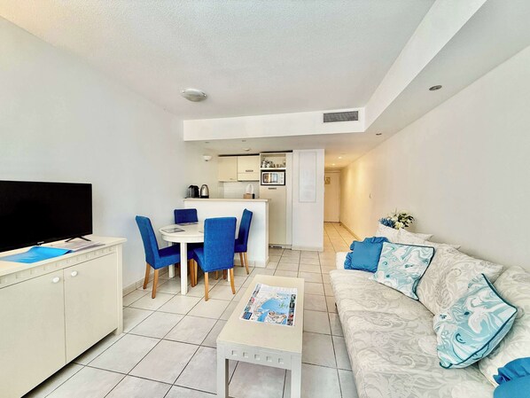 A modern living room with a flat-screen TV, a sofa with patterned cushions, a dining area with blue chairs, and a kitchen in the background.