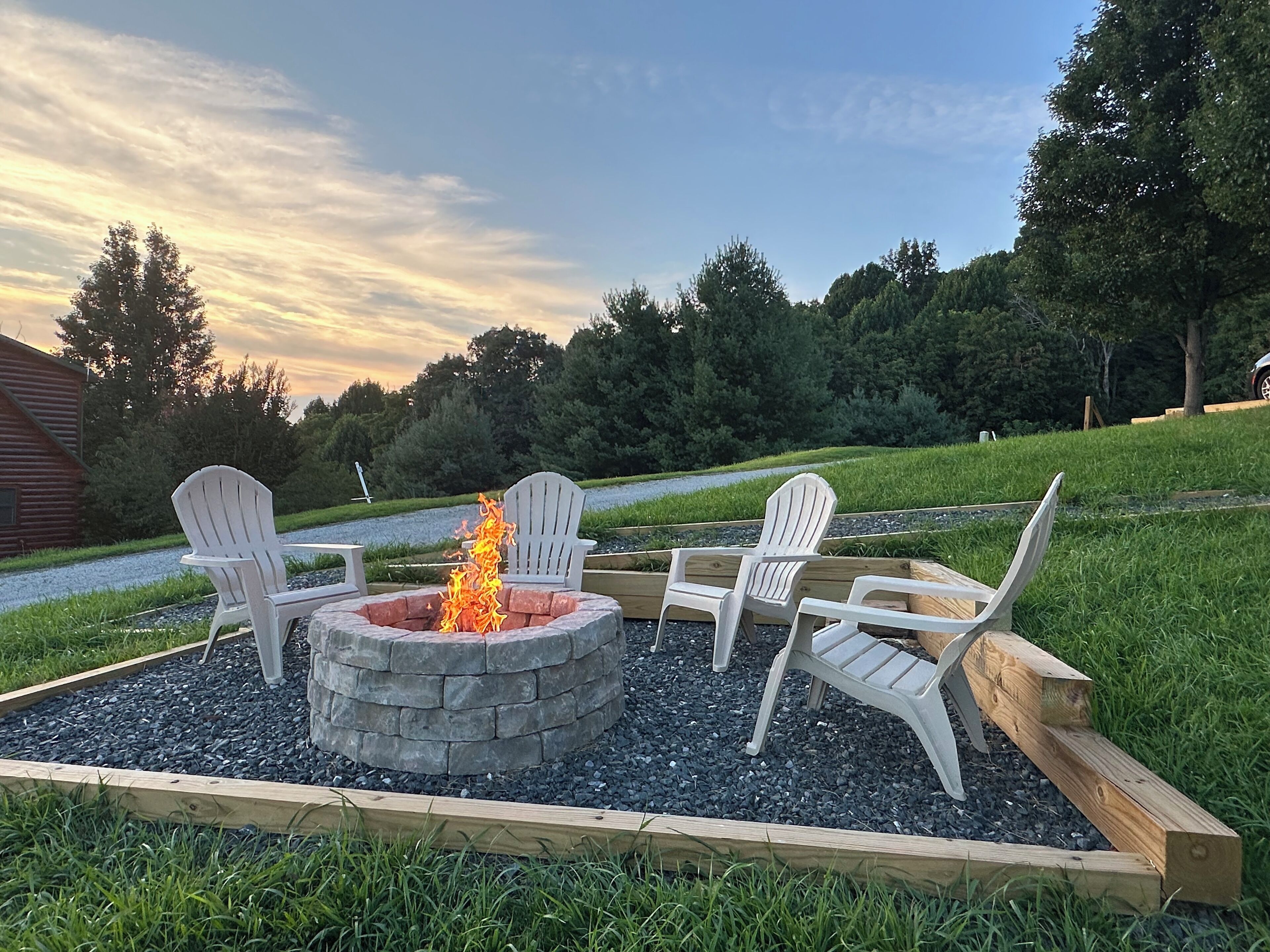 A fire pit with four Adirondack chairs arranged around it, set in a garden with a gravel path and wooden borders.
