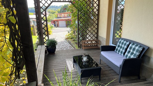 A wooden deck with a sofa and a table, surrounded by potted plants and a lattice structure. - The Property (Rentweinsdorf)