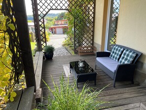 Ett trädäck med en soffa och ett bord, omgivet av krukväxter och en spaljé.