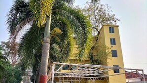 A building with a yellow facade, a covered outdoor area with a metal roof, and palm trees in the background. - Kings Resort (Gobindpur)
