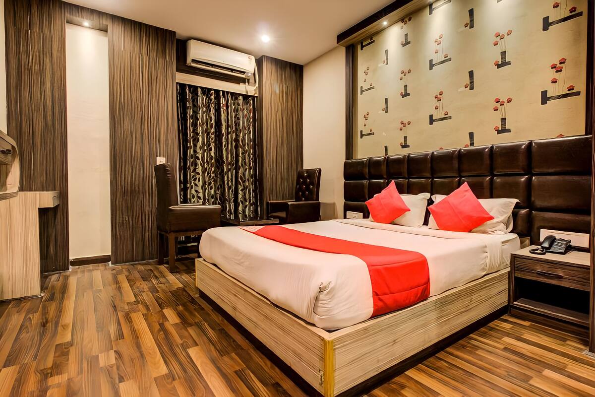 A modern hotel room with a wooden floor, a large bed with red and white bedding, a desk, and a wall-mounted air conditioning unit.