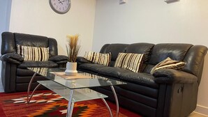 A living room with a glass coffee table, two black leather sofas with striped cushions, a clock on the wall, and a patterned rug. - Crescent Apartment - Two bedroom (Romford)