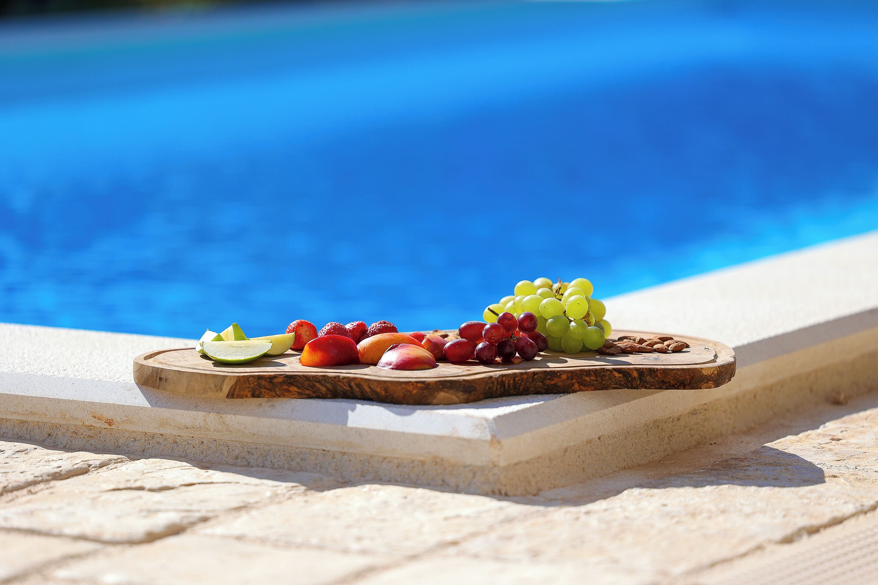 A wooden board with sliced fruits and nuts by a poolside.