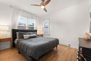 Une chambre à coucher comprenant un lit, des tables de chevet, un ventilateur de plafond et une fenêtre avec des stores.
