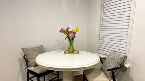 Une table de salle à manger ronde, accompagnée de deux chaises, un vase avec des fleurs et une fenêtre avec des stores.