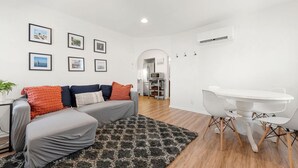 Un salon moderne avec un canapé gris, un tapis noir et blanc, une table à manger blanche avec des chaises, et une cuisine en arrière-plan.