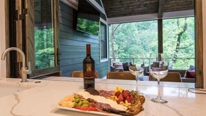 Une table à manger, agrémentée d’une planche de charcuterie, d’une bouteille de vin et de verres, est placée dans une pièce dotée de grandes fenêtres donnant sur une forêt.