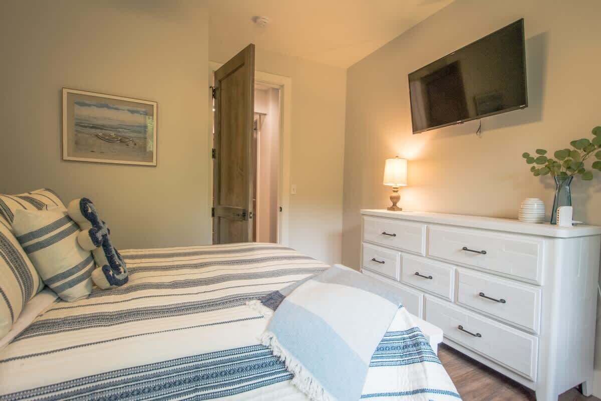 A bedroom with a bed, a TV mounted on the wall, a bedside table with a lamp, and a door leading to another room.