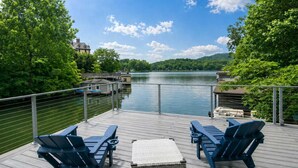 Une terrasse dotée de deux chaises longues et d’un pouf en osier, offrant une vue sur un lac avec un quai et des bâtiments en arrière-plan.