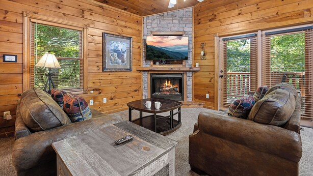L’intérieur chaleureux d’un chalet avec une cheminée, des murs en bois, un ventilateur de plafond et une grande fenêtre donnant sur un paysage verdoyant.