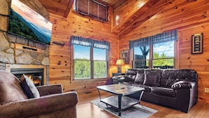 A cozy log cabin interior with a stone fireplace, leather sofa, and a television mounted on the wall. - Views, Hot Tub, Fireplace, Resort Pool, + Game Room (Gatlinburg)