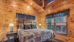 A wooden cabin bedroom with a bed, two nightstands, a lamp, and a window with shutters. - Views, Hot Tub, Fireplace, Resort Pool, + Game Room (Gatlinburg)
