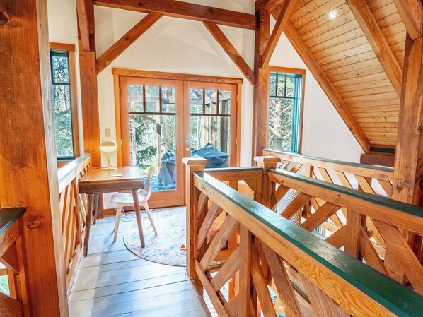 Un interno in legno con una scala che conduce a una zona mansardata, una scrivania con una sedia e ampie finestre con vista sugli alberi.