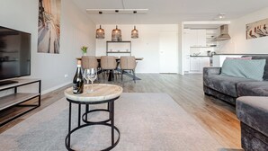 Una sala de estar moderna con televisor, una mesa de centro redonda con una botella y un vaso, un sofá gris y una zona de comedor con mesa y sillas.