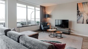 Una sala de estar moderna con un sofá gris, una mesita redonda pequeña, un televisor de pantalla plana y un ventanal grande con cortinas.