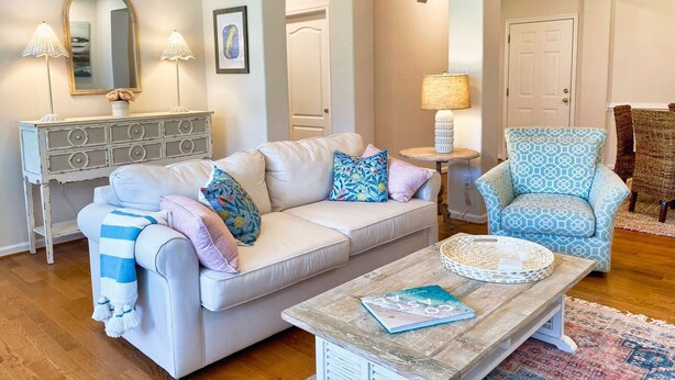 A living room with a white sofa, patterned cushions, a coffee table, a side table with a lamp, a wicker chair, and a mirror on the wall.
