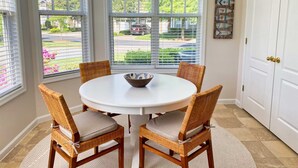 A dining area with a round table, four wicker chairs, a woven pendant light, and a view through large windows.