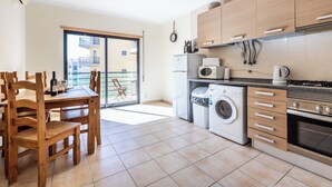 A modern kitchen with a washing machine, oven, and dining area with wooden chairs and a table. - Apartment Lesley - great location! (Armação de Pêra)
