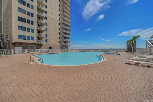 A swimming pool with a brick deck, surrounded by a high-rise building with balconies, and a clear blue sky.