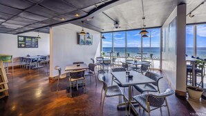 Un área de comedor con mesas y sillas, ventanas amplias con vista a la playa y un panel con la carta.