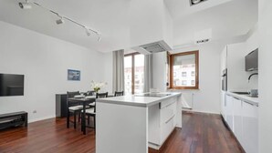 A modern kitchen with a central island, a dining area with chairs, a flat-screen TV, and a window with curtains.