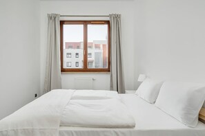 A bedroom with a large window, a bed with white bedding, and a view of buildings outside.