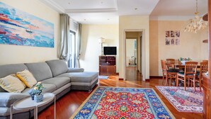 A living room with a grey sofa, a coffee table, a patterned rug, a dining area with wooden chairs and a table, and a painting on the wall. - Elegante Appartamento con Spiaggia sul Mare di Bari (Bari)