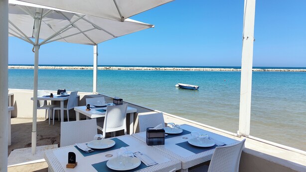 A seaside dining area with white wicker furniture, a table set for two, and a view of the sea and a boat in the distance. - Elegante Appartamento con Spiaggia sul Mare di Bari (Bari)
