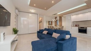 Una sala de estar moderna con un sofá seccional azul, un televisor de pantalla plana montado en la pared y una cocina al fondo.