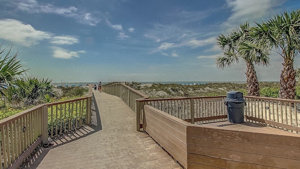 Ein hölzerner Steg führt zu einem Strand mit Palmen und blauem Himmel.
