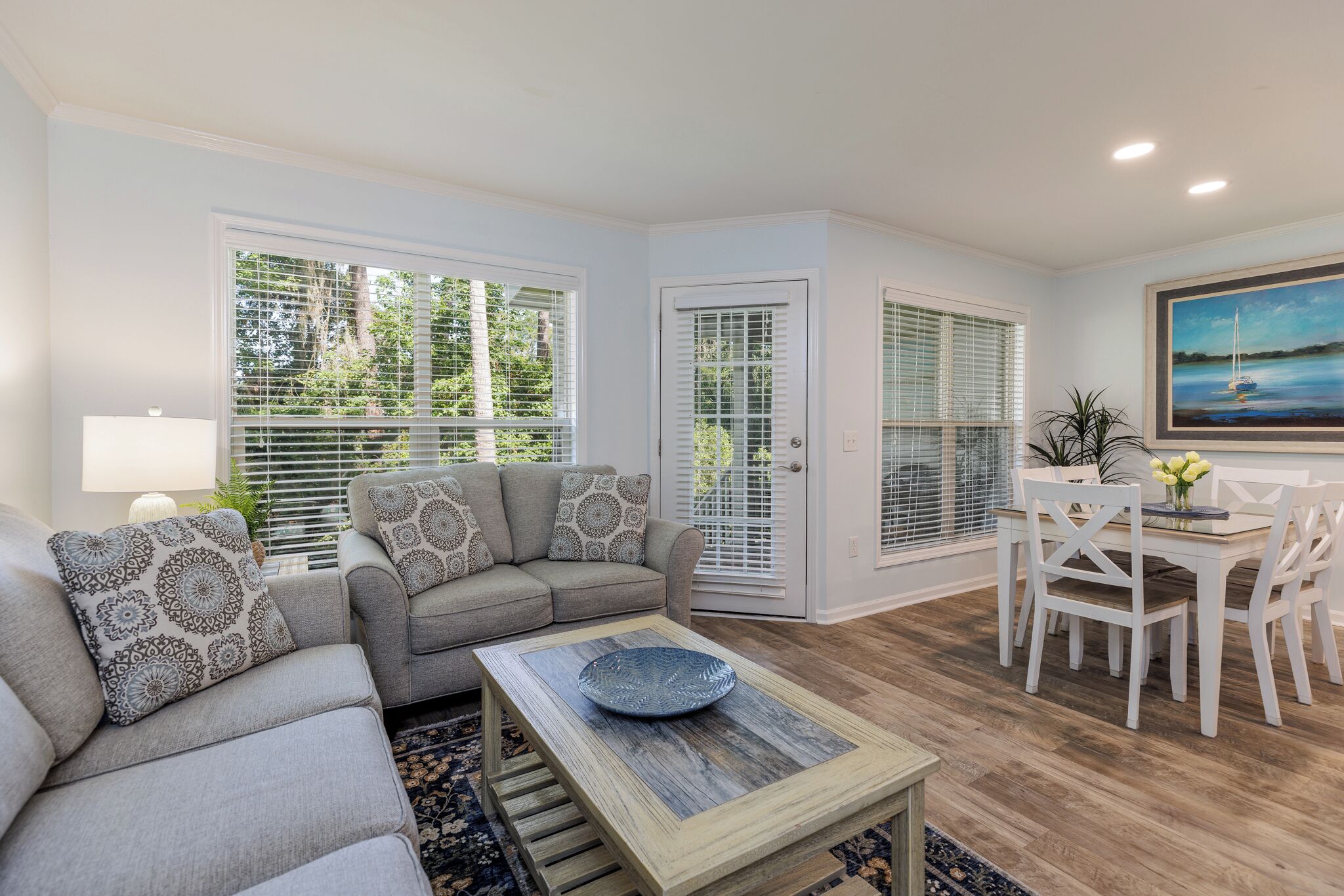 A living room with a grey sofa, patterned cushions, a wooden coffee table, and a dining area with white chairs and a table.
