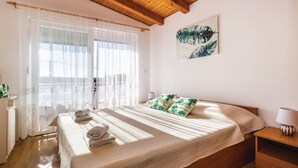 A bedroom with a wooden ceiling, a large window with sheer curtains, a bed with a quilt and pillows, and a painting on the wall.