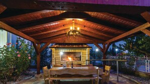 An outdoor patio with a stone floor, wooden table, and chairs, featuring a stone fireplace under a wooden pergola.