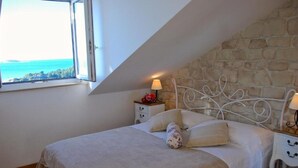 A bedroom with a stone wall, a bed with white bedding, two bedside tables with lamps, and a window with a view of the sea. - Villa Mediterranea (Zupa dubrovacka)