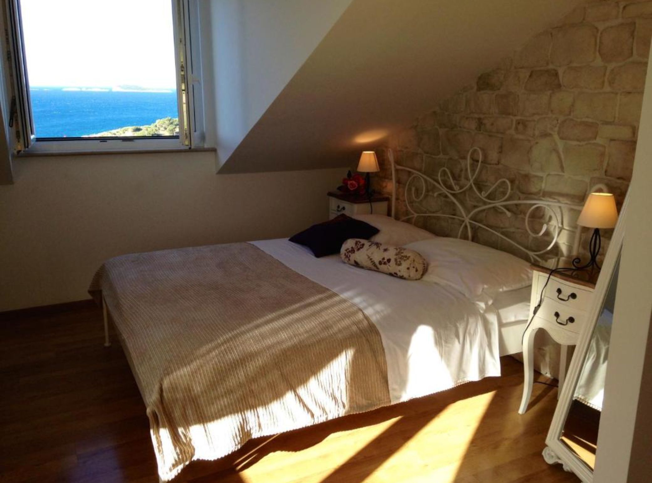 A bedroom with a stone wall, a bed with a white headboard, a nightstand with a lamp, and a view of the sea through a window.