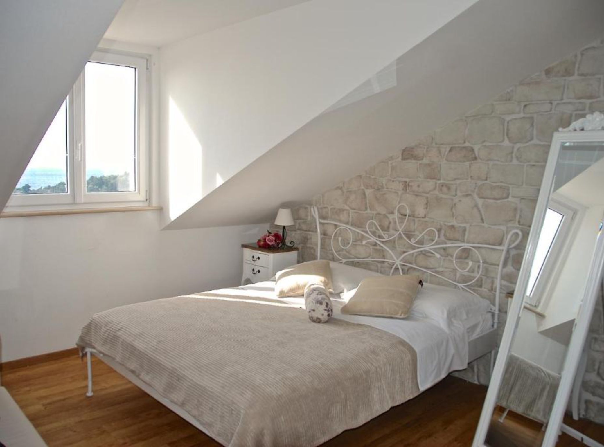 Une chambre avec un mur en pierre, un lit avec un cadre en métal blanc, une table de chevet avec une lampe et une fenêtre donnant sur un beau paysage.