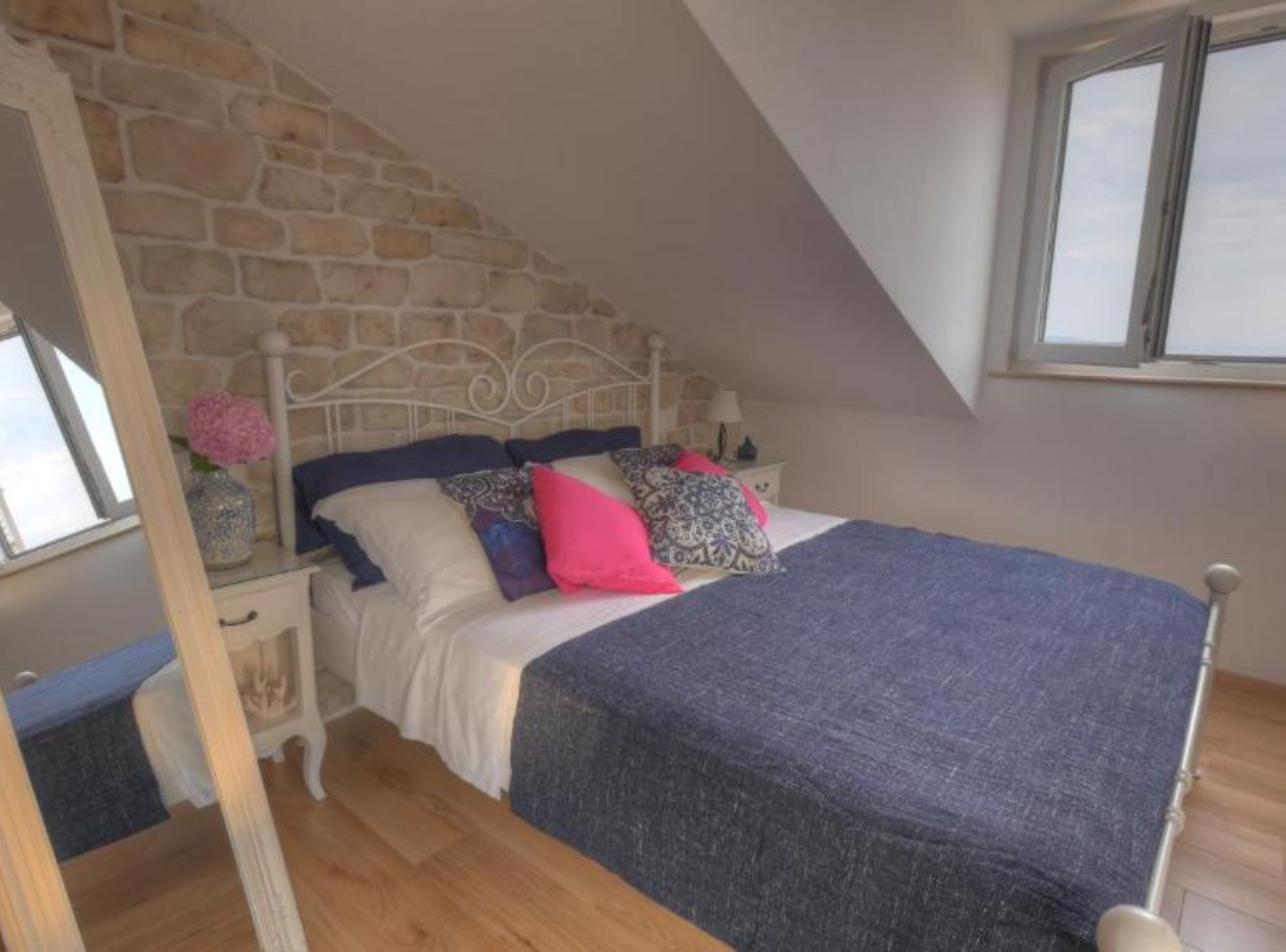 A bedroom with a stone wall, a bed with multiple pillows, a nightstand, a lamp, and a window.