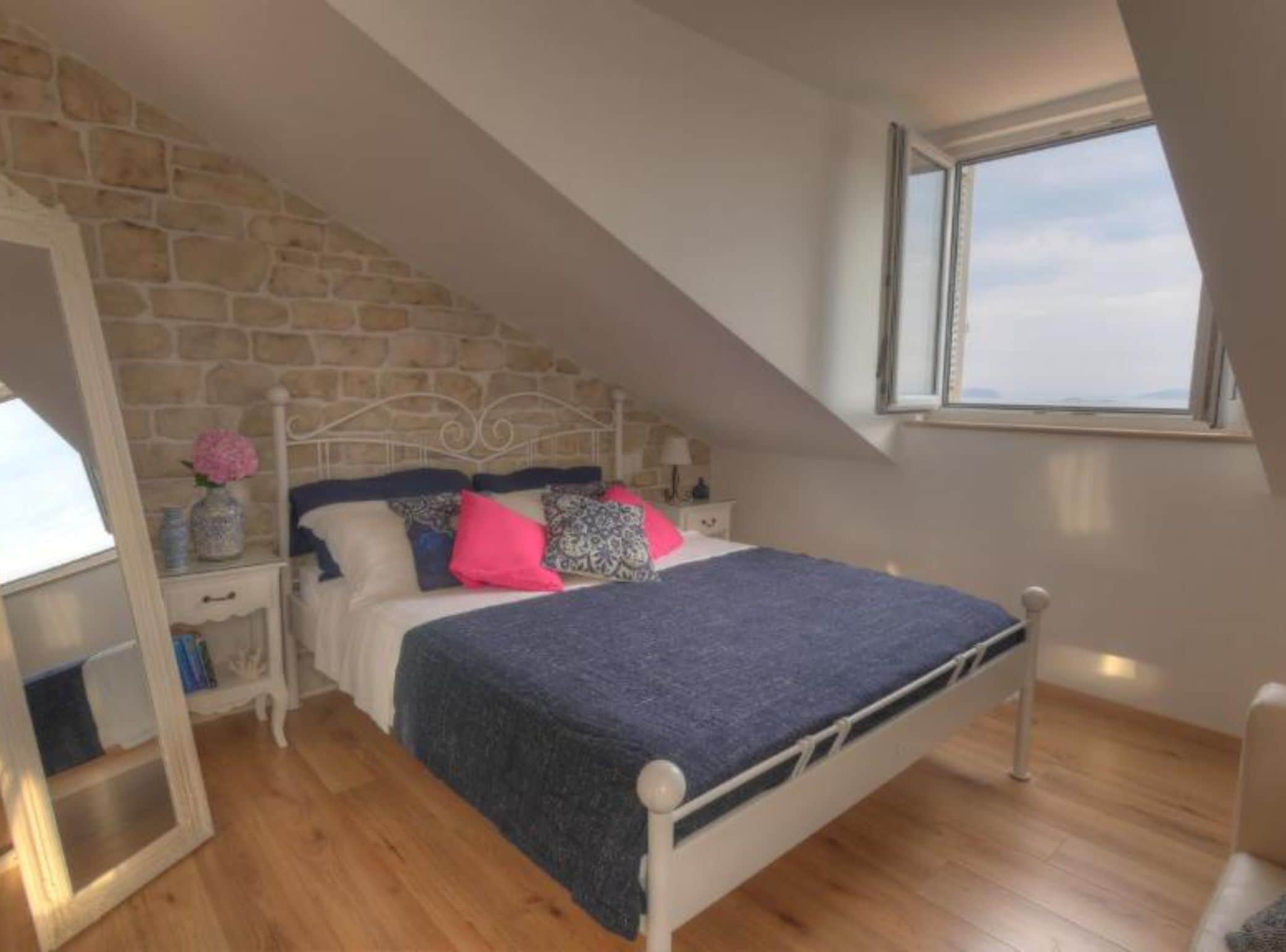 A bedroom with a stone wall, a bed with a blue blanket, and a window with a view.