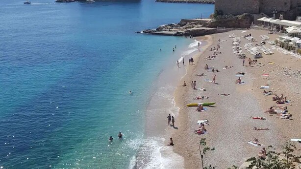 A beach with clear blue waters, a historic building complex in the background, and a sandy shore with umbrellas and people. - Apartment Ivančica (Dubrovnik)
