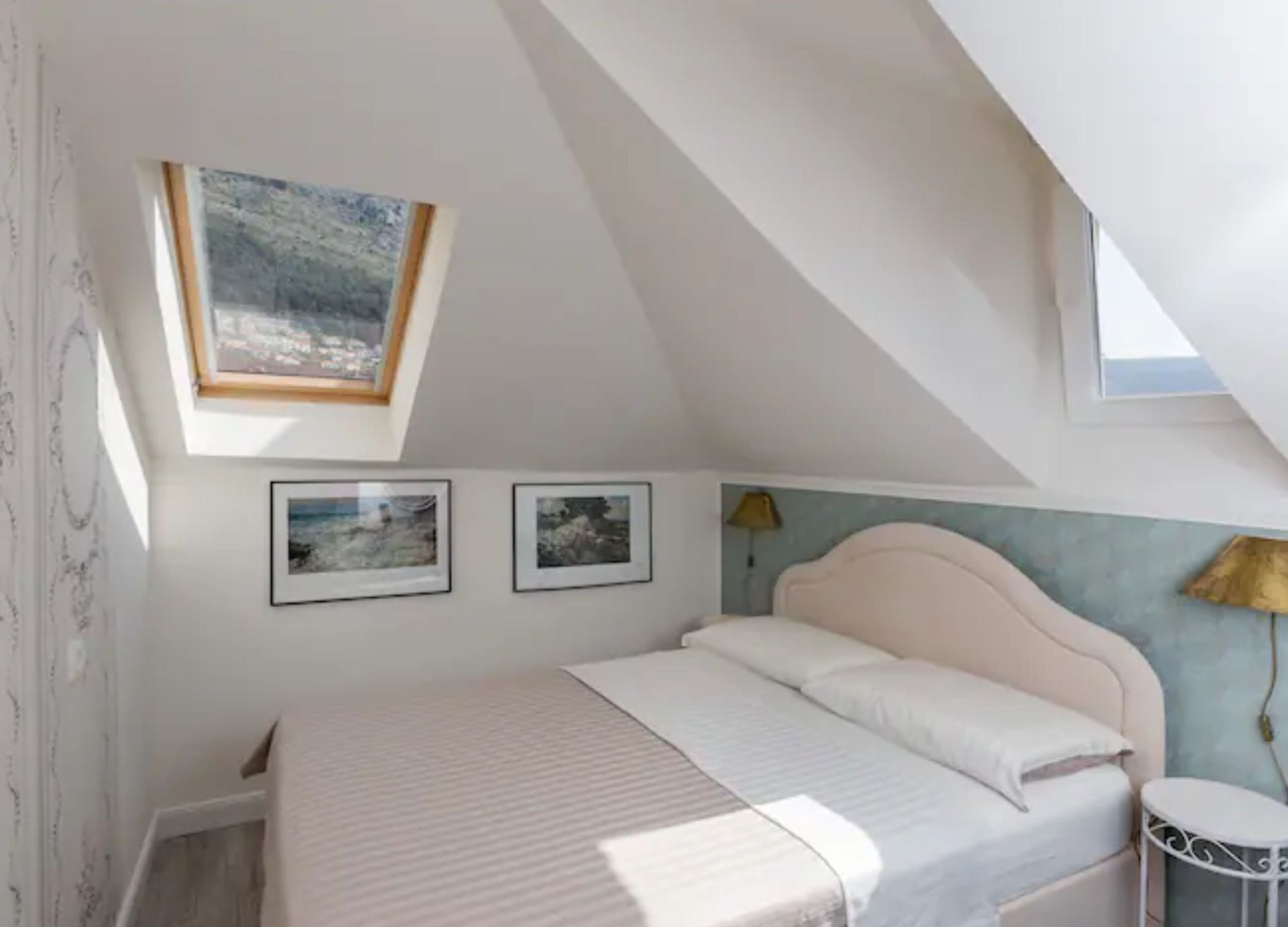 Ein Schlafzimmer mit schrägen Wänden, einem Bett mit weißer Bettwäsche, zwei gerahmten Bildern an der Wand, einem kleinen runden Tisch und einer Lampe.
