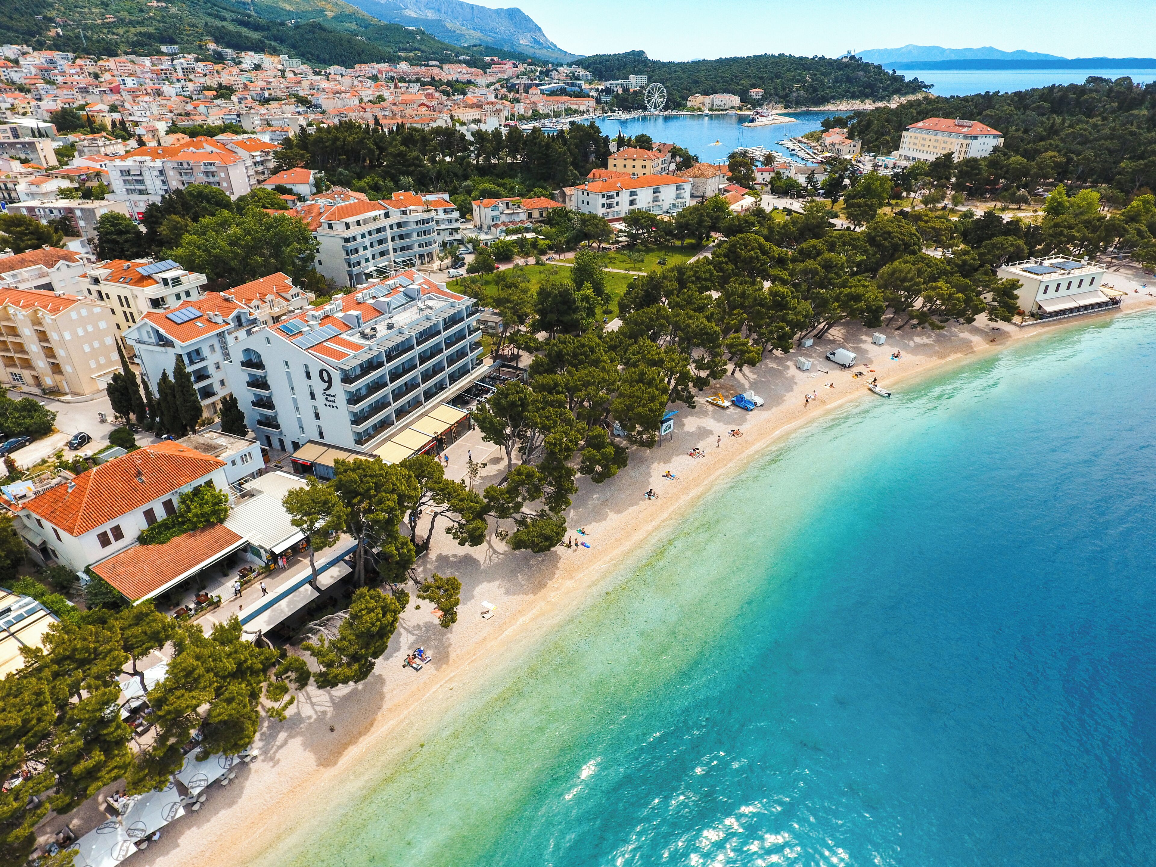 Una vista aerea di una località costiera con una spiaggia, edifici con tetti in tegole rosse e un mare blu cristallino.