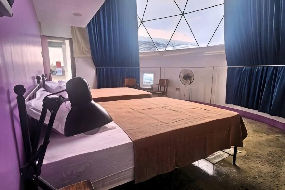 Ett rum med två sängar, ett skrivbord, en stol, ett fönster med gardiner och en fläkt.