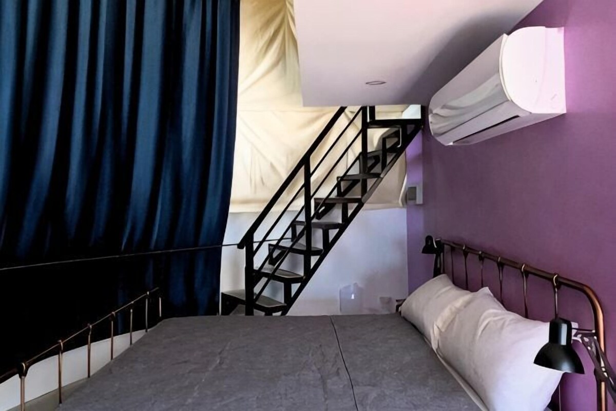 A bedroom with a bed, a black metal headboard, a black lamp, a white air conditioning unit, a staircase, and a purple wall.